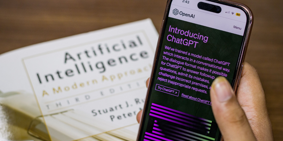 En telefon öppen med OpenAIs ChatGPT uppe på skärmen och en kompendium om artificiell intelligens i bakgrunden på ett träbord.
